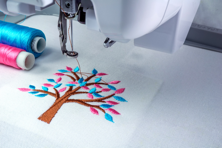Nahaufnahme einer Stickmaschine beim Sticken eines Baummotivs mit braunem Stamm und pinken sowie türkisfarbenen Blättern auf weißem Stoff; daneben liegen passende Garnrollen.